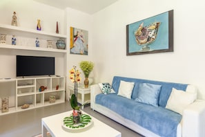A living room with a white sofa, a flat-screen TV, decorative items on shelves, and a painting on the wall.
