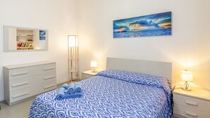 Een slaapkamer met een bed, nachtkastjes, een dressoir, een boekenkast en een schilderij van een zonsondergang boven de oceaan.