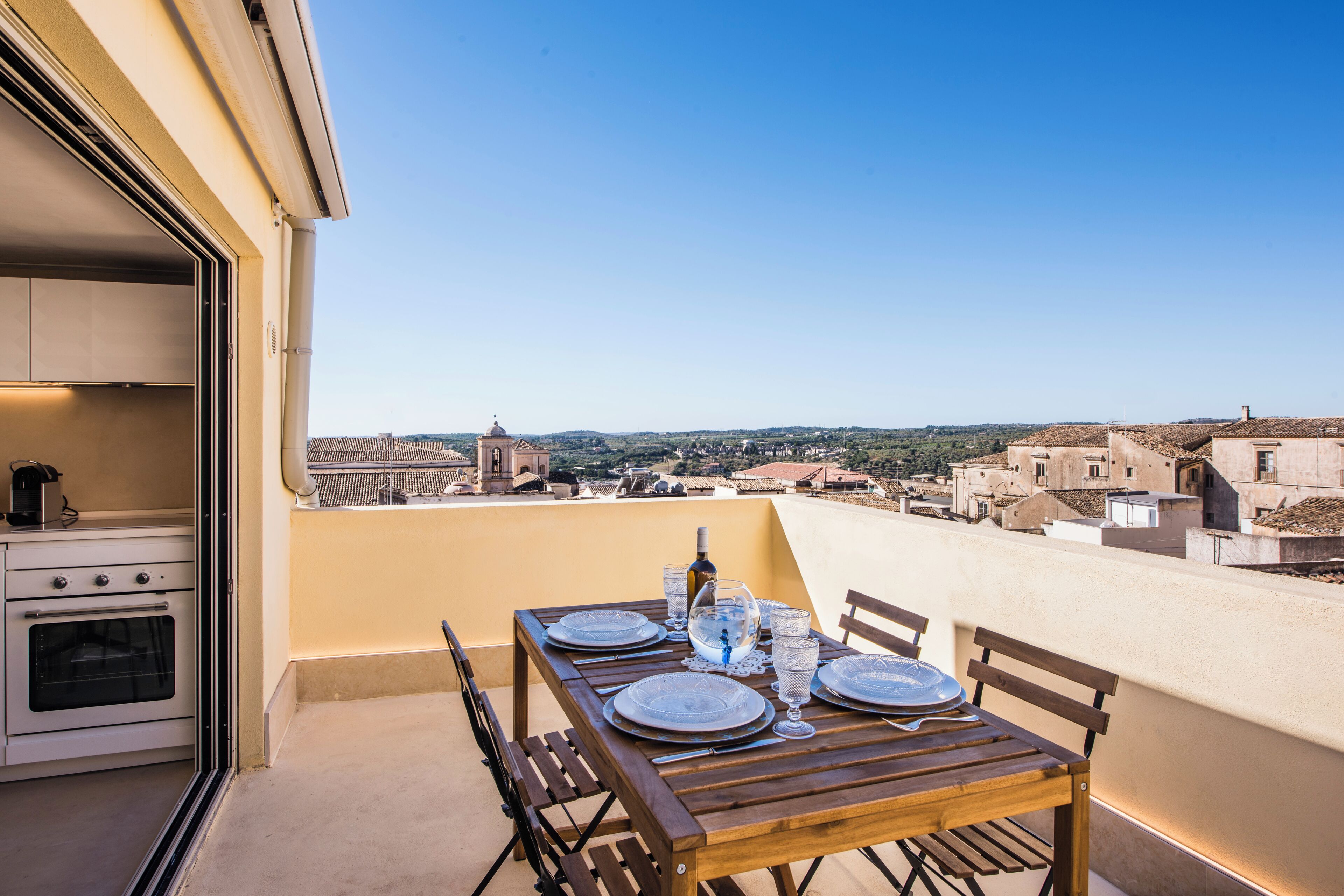 A balcony with a wooden table set for dining, overlooking a cityscape with historical buildings.