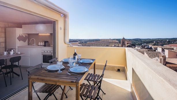 A balcony with a wooden table set for two, overlooking a cityscape with historical buildings. - Casa Novella con Terrazza by Wonderful Italy (Noto)