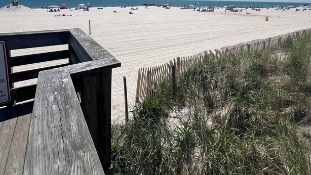 Una playa con un camino de madera que lleva a la orilla, arena y cielo azul despejado.