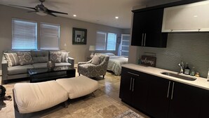 Una sala de estar moderna con un sofá, un sillón y una mesa de comedor. Hay una zona de cocina con armarios oscuros y una encimera.