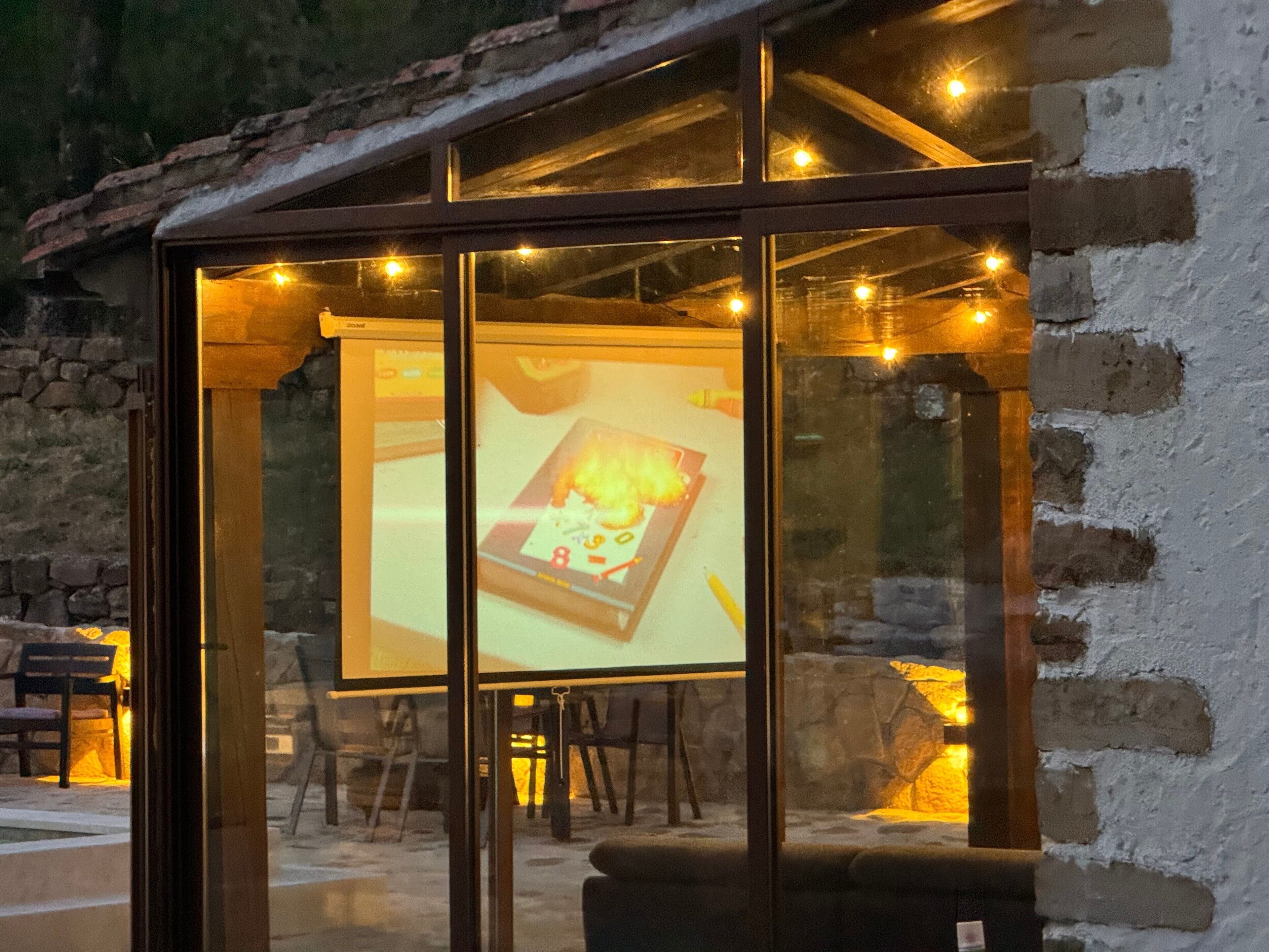 A glass-walled room with a projection screen displaying a fire, stone walls, and string lights.