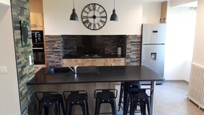 A modern kitchen with a central island, black bar stools, a large wall clock, and a refrigerator. - Holiday Home 'Gîte La Chartrouille' with Private Terrace, Private Garden and Wi-Fi (Allassac)