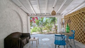 Une terrasse couverte avec un sol en pierre, un canapé, une petite table et des chaises bleues.