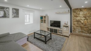 A modern living room with a sofa, a glass coffee table, a TV, a bookshelf, and a stone wall. - Casa La Molina – Spacious Countryside Home with Mountain Views near Burgos (Burgos)