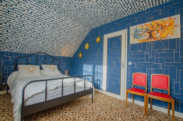 A bedroom with a patterned blue and white ceiling, a bed with white bedding, two red chairs, and a door with a geometric pattern.