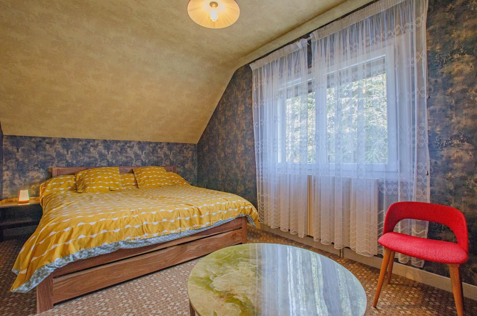 A bedroom with a bed, a chair, a small table, and a window with sheer curtains.