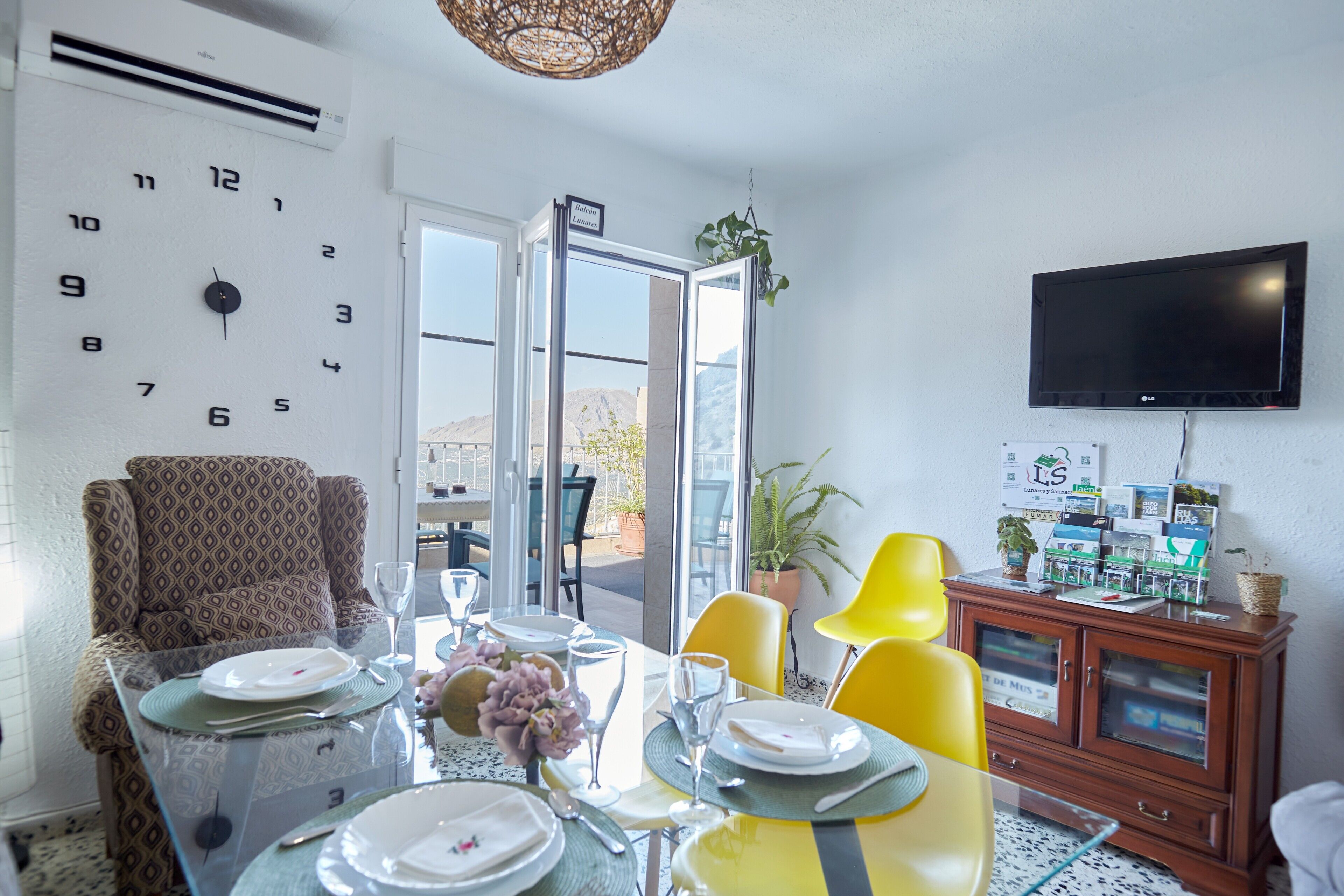 A dining area with a table set for four, a clock on the wall, and a television mounted above a cabinet.