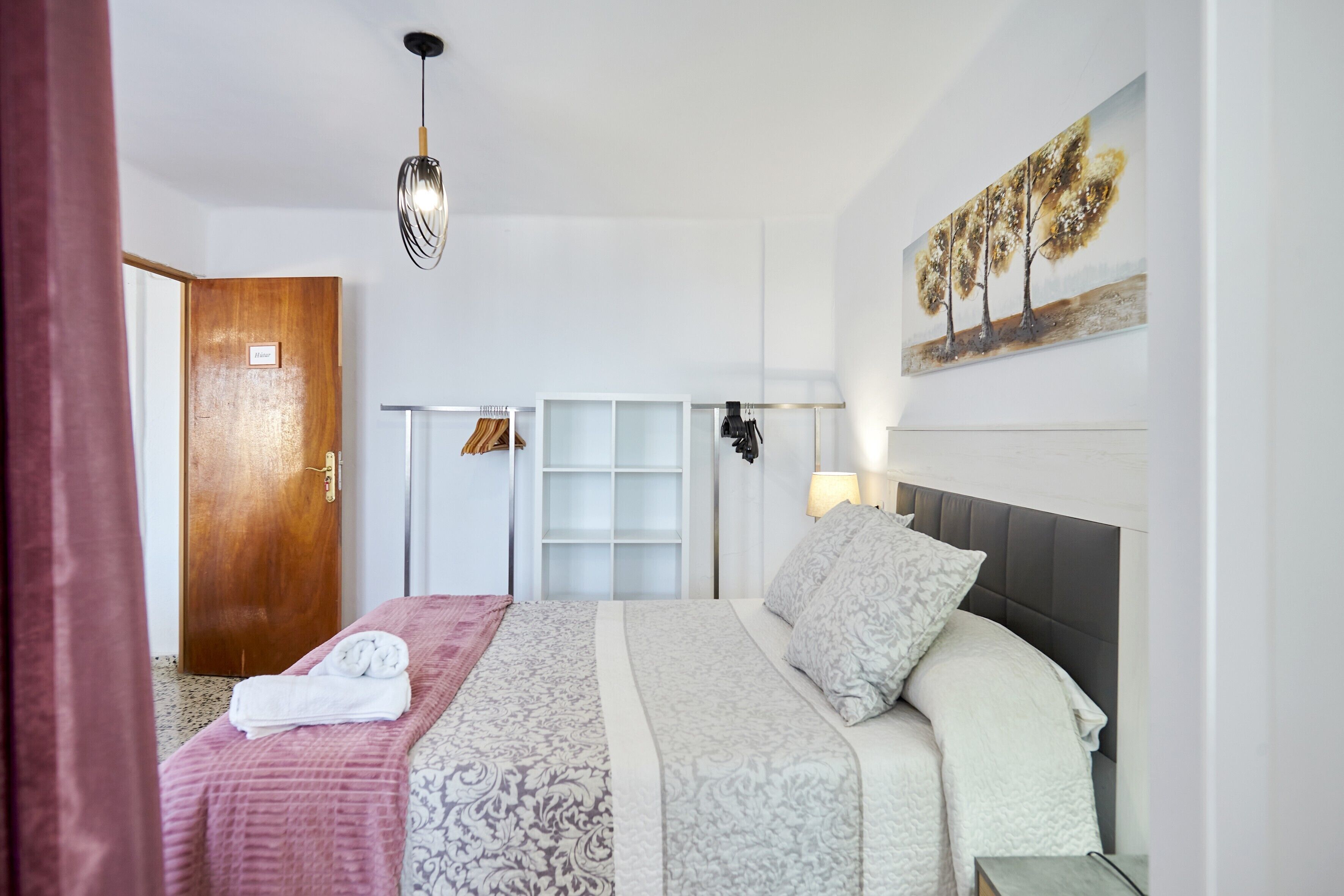 Una habitación de hotel con cama, una puerta de madera, un armario, un cuadro y una lámpara colgante.