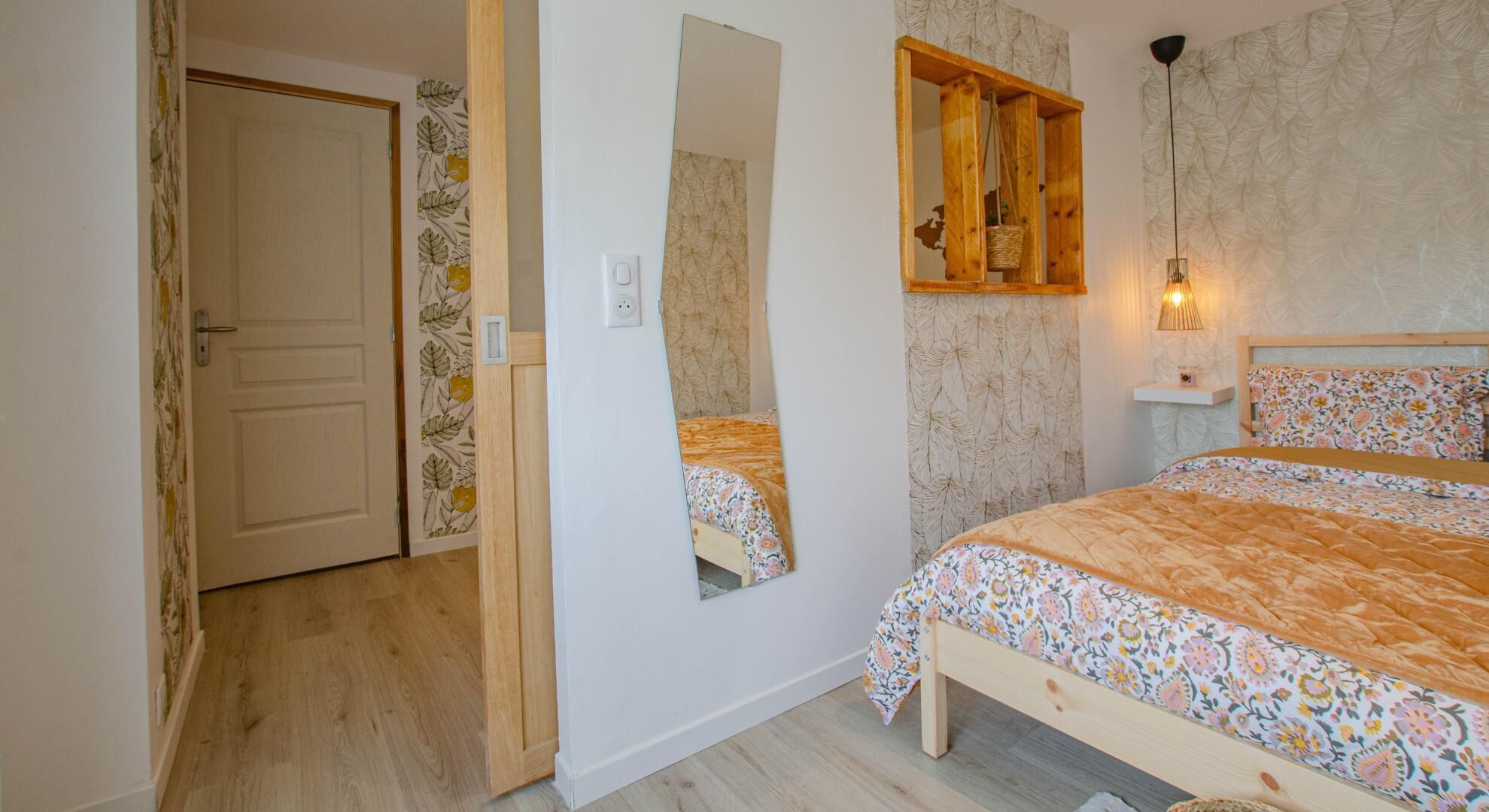 Ein Schlafzimmer mit einem hölzernen Bett, einem Spiegel, einer Pendelleuchte und einer Tür mit floralem Wandpapier.