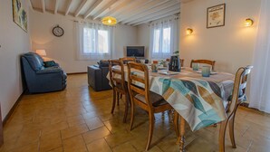 A dining area with a wooden table, chairs, and a patterned tablecloth. There is a sofa and a television in the background. - Le Marguette - PEPPERS (POIVRES)