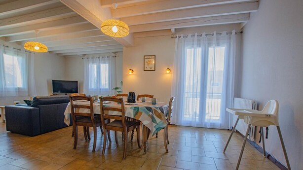 A dining area with a table, chairs, and a high chair. There is a sofa and a television in the background. - Le Marguette - PEPPERS (POIVRES)
