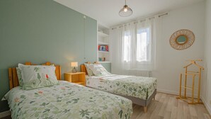 A bedroom with two beds, a wooden nightstand, a hanging light fixture, a radiator, and a wooden hanger rack. - Le Marguette - PEPPERS (POIVRES)