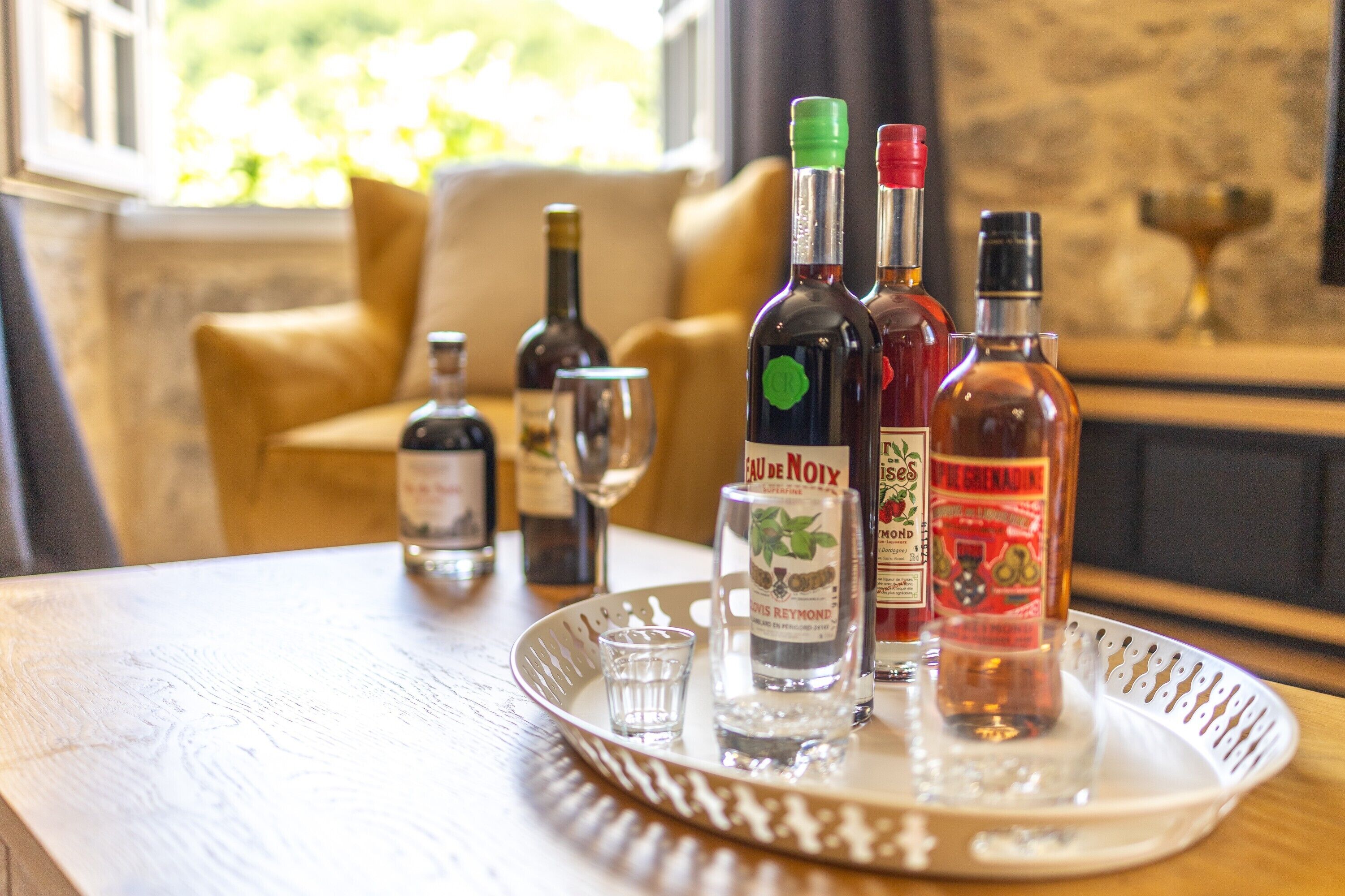 Una bandeja con varias botellas de licor y una copa para shots sobre una mesa de madera en una habitación con un sofá beige y una ventana con vista a follaje.
