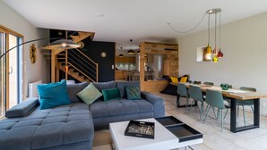 Una sala de estar moderna con un sofá gris, una zona de comedor con sillas y una mesa, una cocina al fondo y una escalera que sube a un nivel superior.