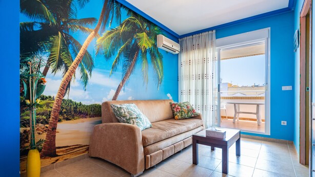 A living room with a sofa, a small table, a large mural of palm trees, and a view through a glass door.