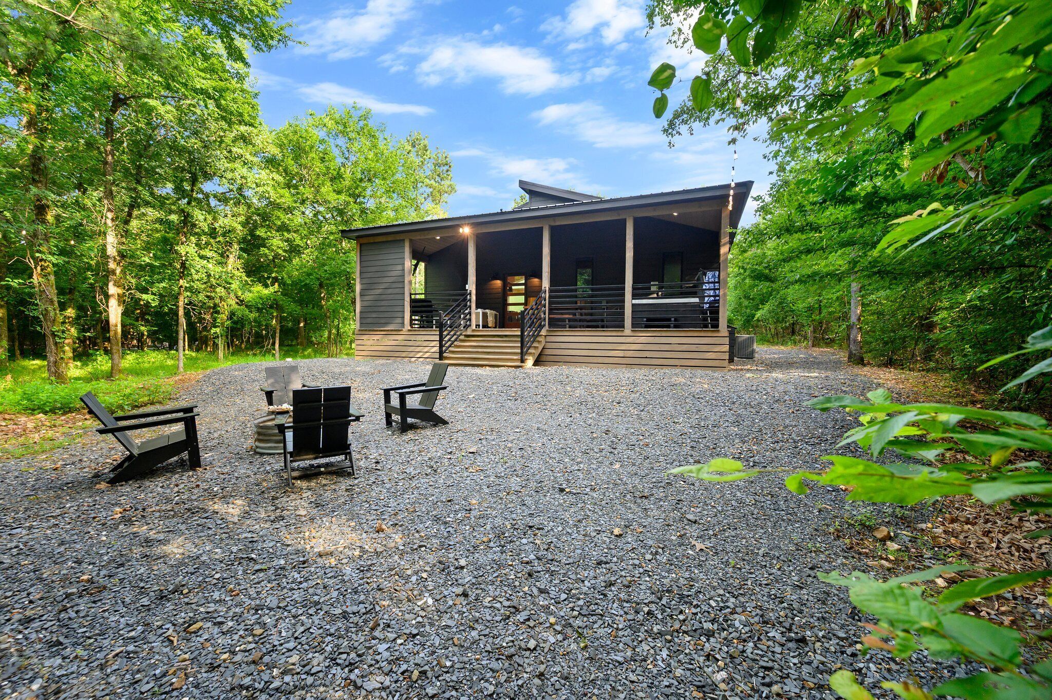 A modern cabin with a covered porch, outdoor seating, and a gravel driveway surrounded by trees.
