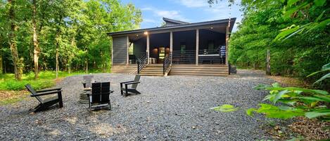 A modern cabin with a covered porch, outdoor seating, and a gravel driveway surrounded by trees.