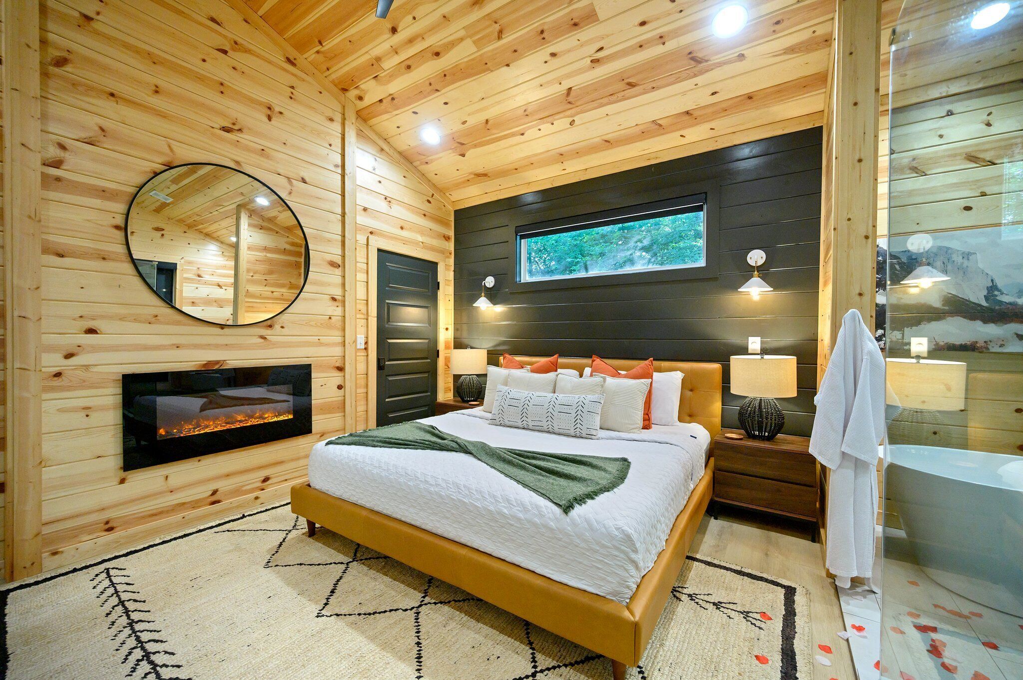 A wooden-paneled bedroom with a bed, bedside lamps, a mirror, a fireplace, and a large window.