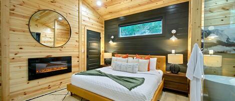 A wooden-paneled bedroom with a bed, bedside lamps, a mirror, a fireplace, and a large window.