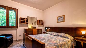 Ein Schlafzimmer mit einem Holzbett, einem Schminktisch, einem Spiegel, einem Nachttisch mit Lampe und einem Fenster mit Blick auf Grünflächen.