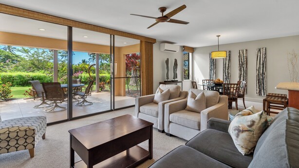 A spacious living area with a sofa, armchairs, and a coffee table. There is a ceiling fan and a dining table with chairs. The room has large windows offering a view of a garden.