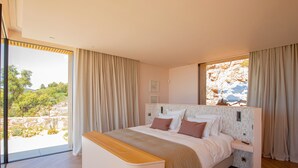 A modern bedroom with a large bed, wooden headboard, and a view of a rocky landscape through a large window. - Villa Lucia Di Arinella (Zonza)