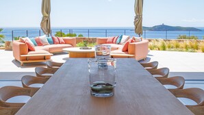 A modern outdoor seating area with a wooden table, cushioned sofas, and a view of the sea. - Villa Lucia Di Arinella (Zonza)