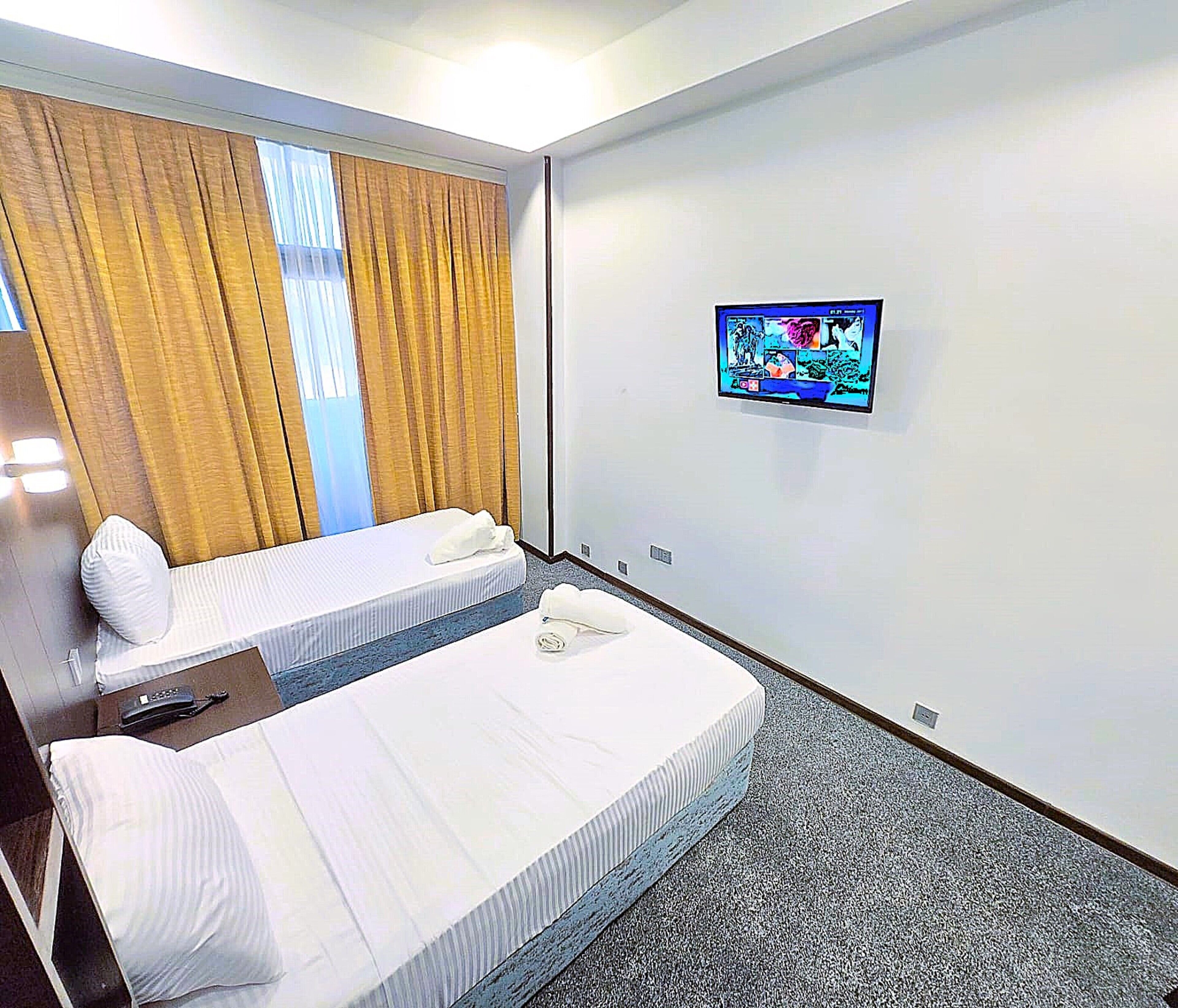 A hotel room with two beds, a television on the wall, and a window with curtains.
