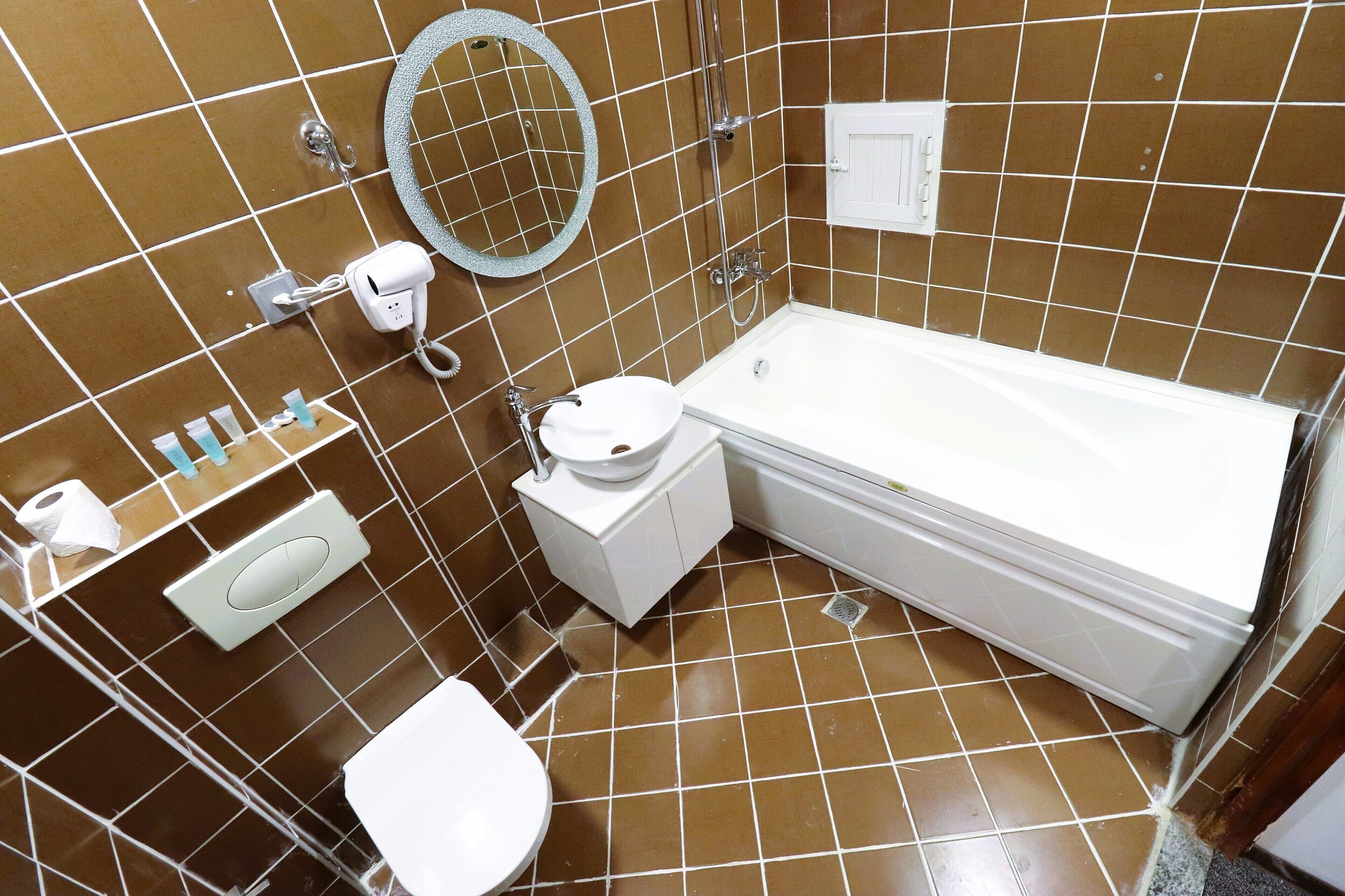 Une salle de bain avec des carreaux marron, des toilettes blanches, un lavabo, un miroir et une baignoire.