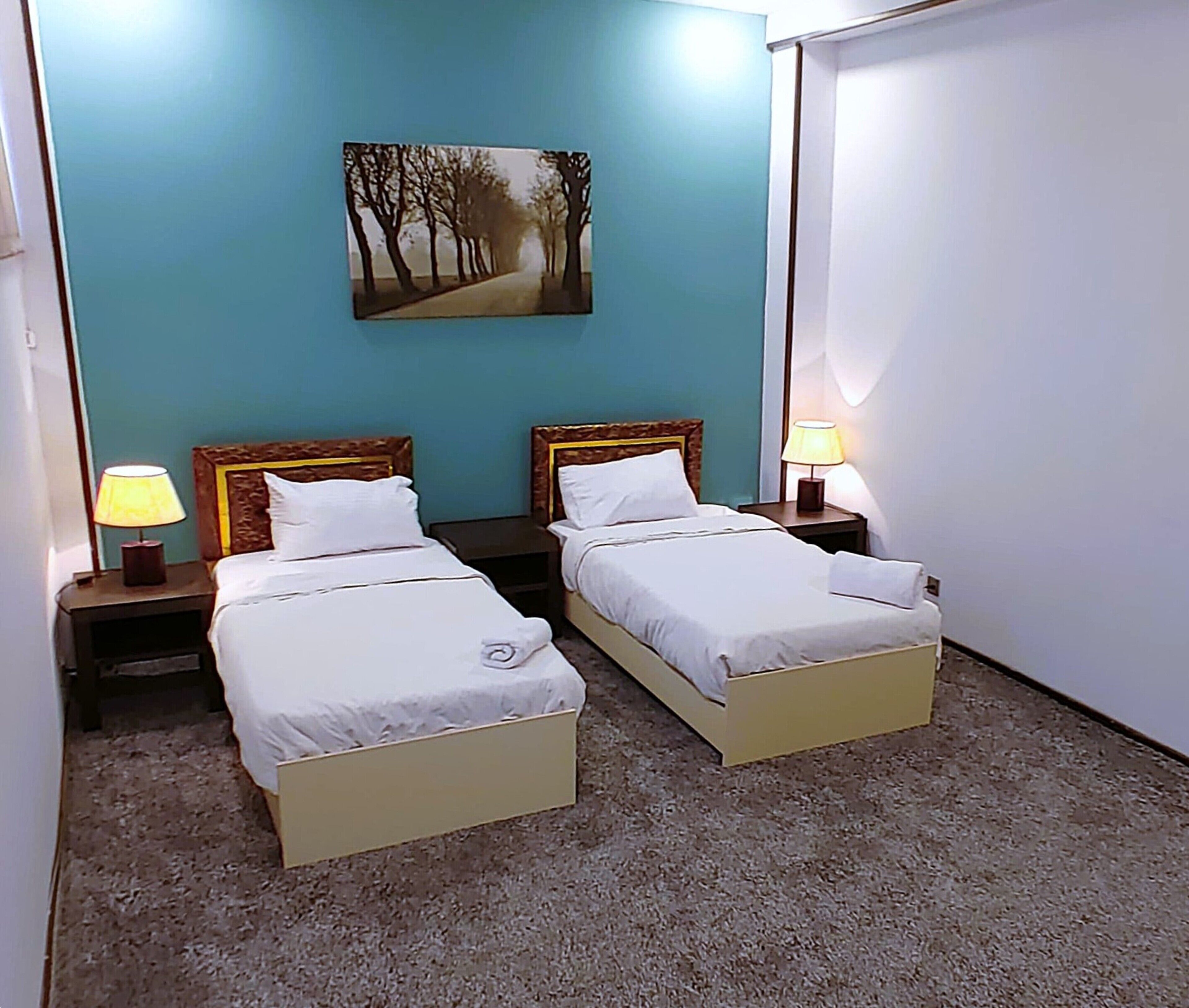 A hotel room with two single beds, a painting of a tree-lined path on the wall, and a carpeted floor.