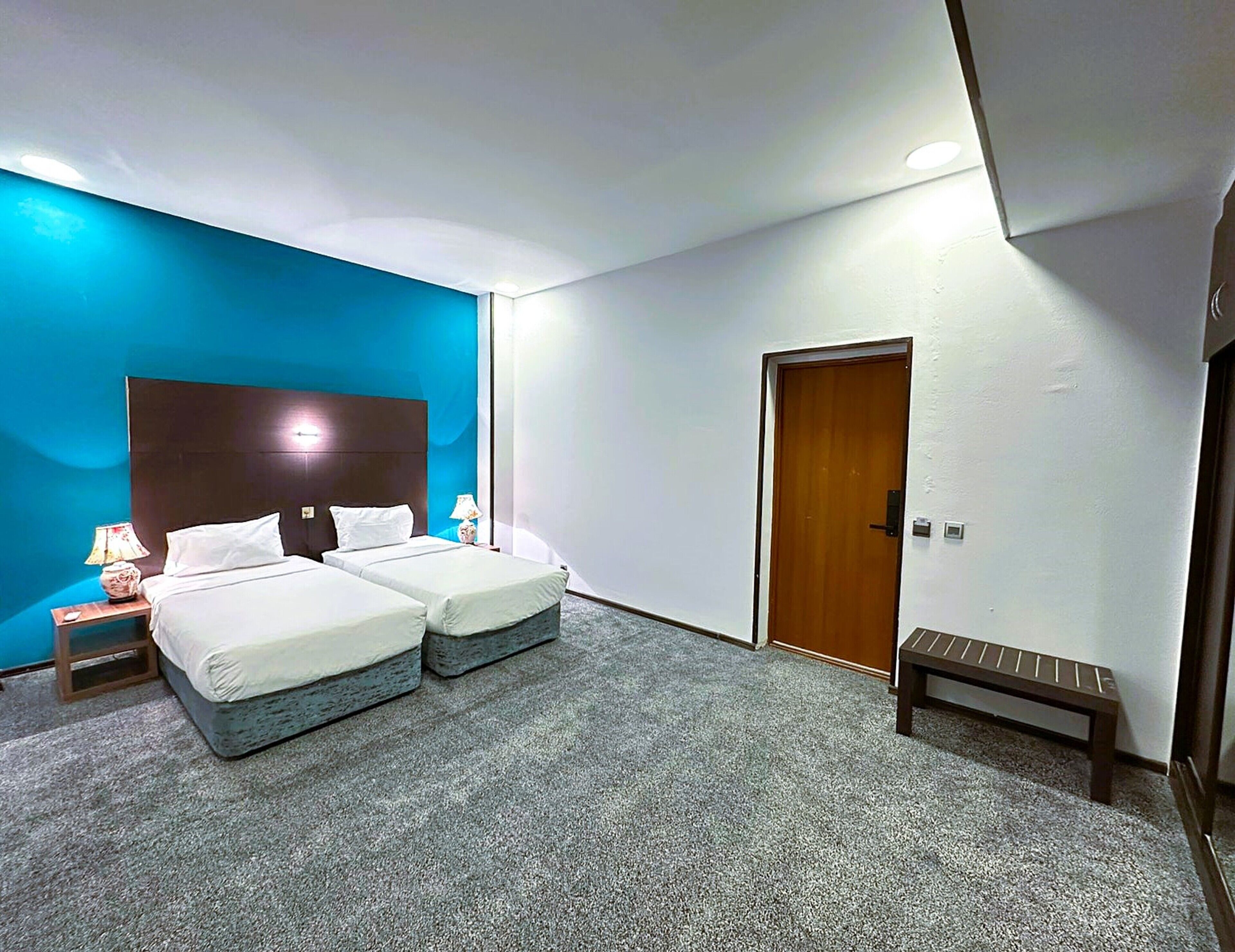 A hotel room with two beds, a bench, a nightstand with a lamp, and a door.