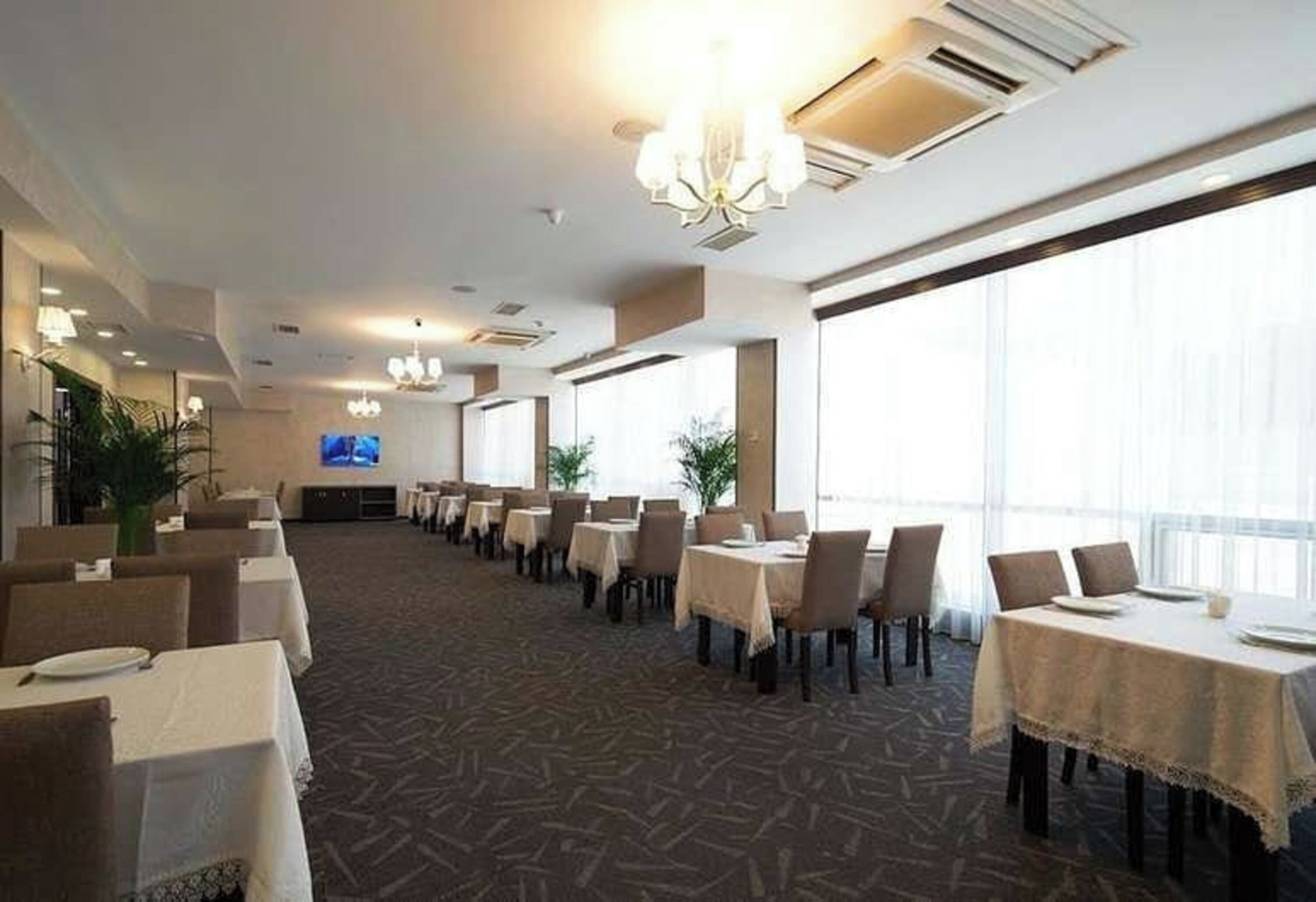 A hotel dining area with multiple tables and chairs, large windows, and a chandelier.
