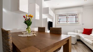 A modern dining area with a wooden table, wicker chairs, a vase of flowers, and a white sofa with a red pillow.