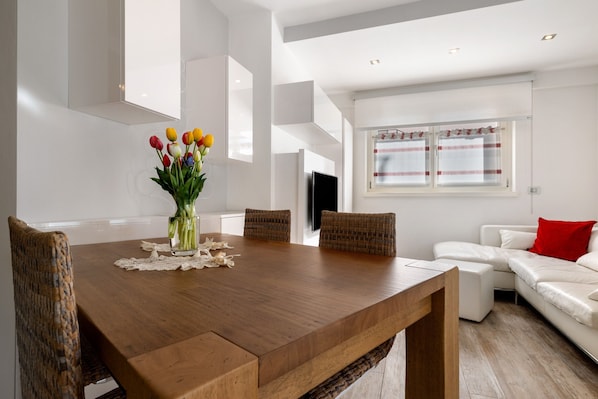 A modern dining area with a wooden table, wicker chairs, a vase of flowers, and a white sofa with a red pillow.
