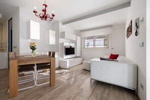 A modern living room with a dining area, a white sofa, a TV, and a chandelier.