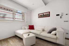 A modern living room with a white sectional sofa, a small white ottoman, a red pillow, a framed picture on the wall, and a window with curtains.
