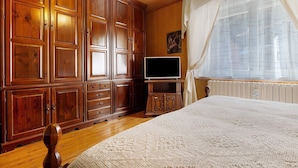 Ein Schlafzimmer mit Holzausstattung, einem Fernseher auf einem Standfuß, einem Bett mit weißer Bettdecke und einem Fenster mit durchsichtigen Vorhängen.