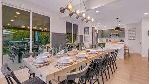 Un espace de repas moderne avec une longue table dressée pour un repas, une cuisine en arrière-plan et de grandes fenêtres offrant une vue sur l’extérieur.