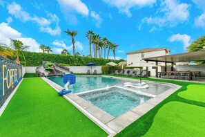 Une piscine avec une pelouse artificielle, un toboggan et un bâtiment doté d’une terrasse.