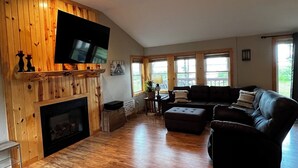 A spacious living room with a fireplace, a large flat-screen TV mounted on a wooden wall, a sectional sofa, and large windows providing natural light. - Heaven on Lake Edward  (Nisswa)