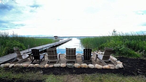 A wooden deck with Adirondack chairs facing a calm lake, a dock extending into the water, and a grassy shoreline. - Heaven on Lake Edward  (Nisswa)