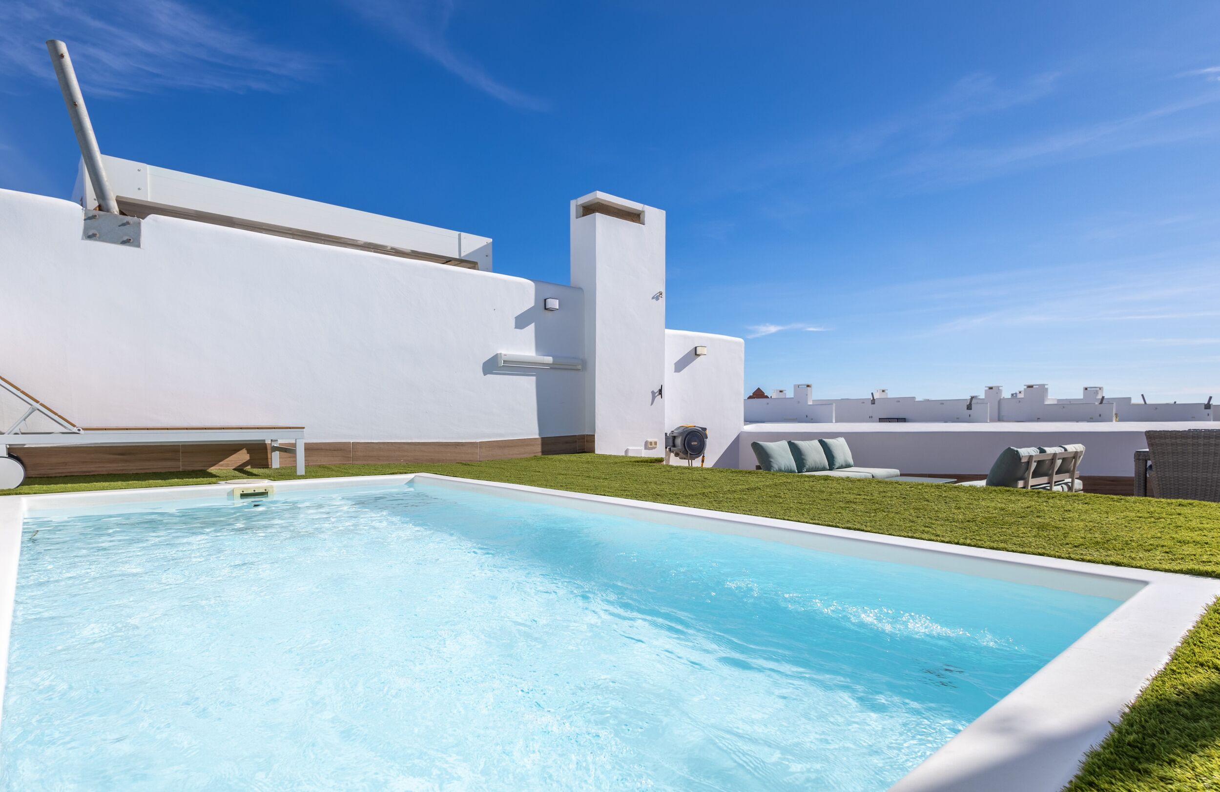 A rooftop pool with a clear blue sky, white walls, and a grassy area with lounge chairs.
