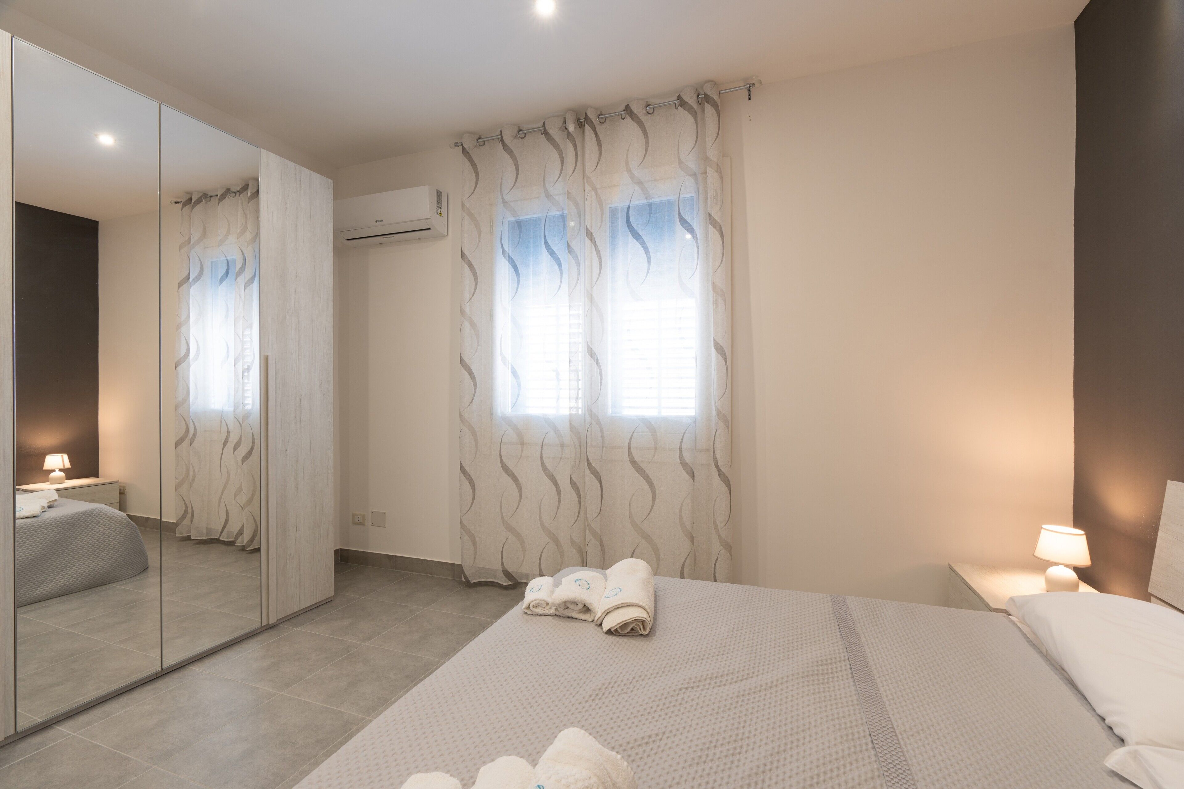 A hotel room with a large mirror, a bed with white linens, a bedside table with a lamp, and a window with curtains.