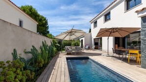 En swimmingpool med træterrasse, udendørs møbler og parasoller.