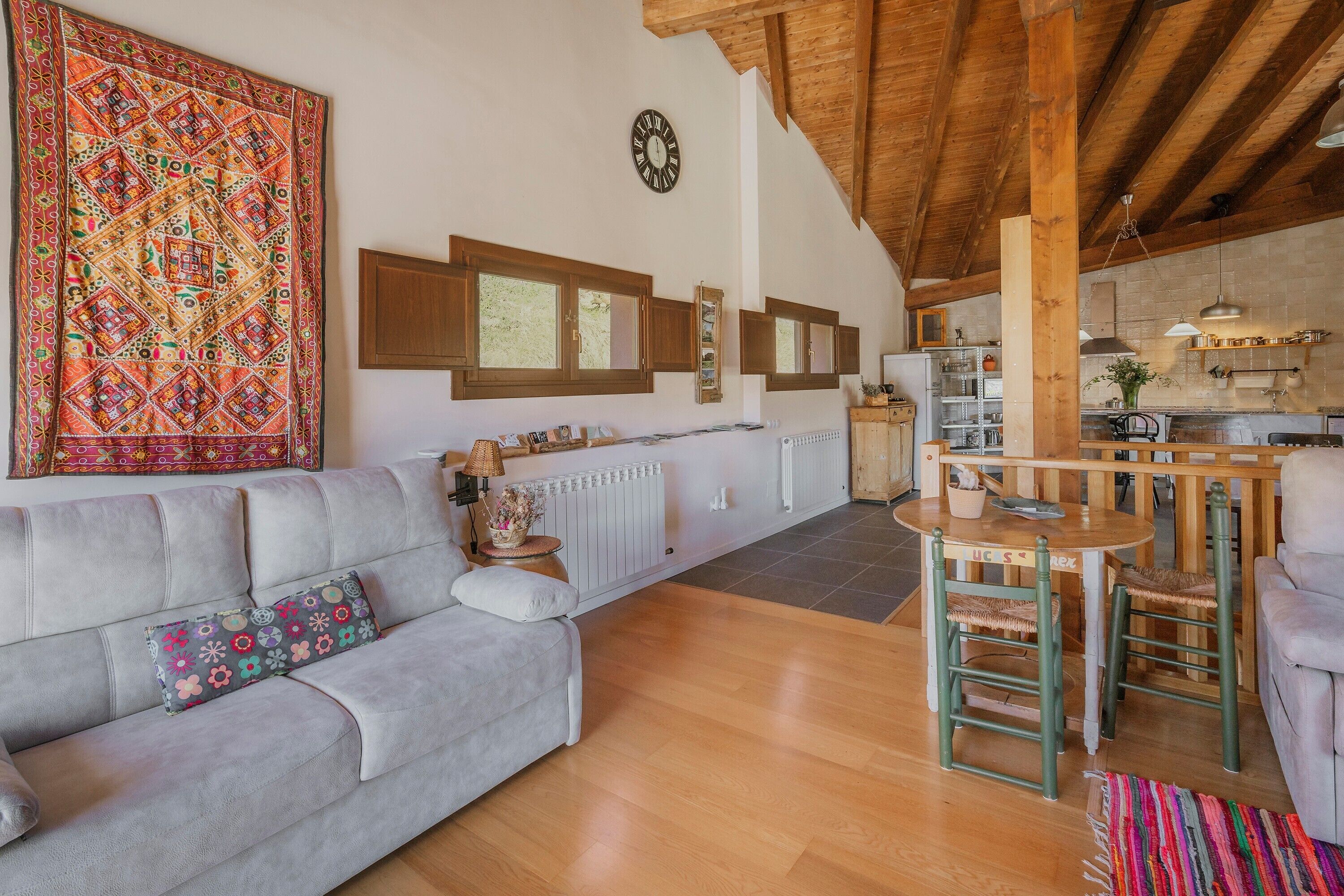 A cozy living room with a sofa, a dining area with a table and chairs, a kitchen in the background, and a large decorative tapestry.