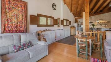 A cozy living room with a sofa, a dining area with a table and chairs, a kitchen in the background, and a large decorative tapestry.