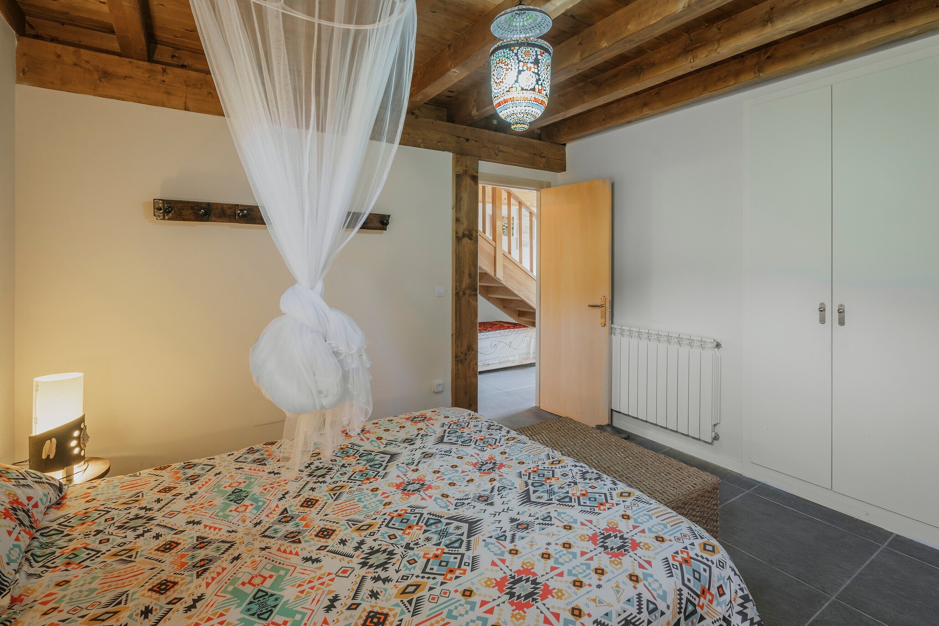 Une chambre à coucher comprenant un lit, une table de chevet, une lampe de chevet, un lit à baldaquin, une couvre-lit à motifs, un plafond en bois, un lustre, une armoire blanche, une porte blanche et un escalier.