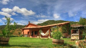 Une maison de campagne au toit en tuiles, avec un hamac et une pompe à eau, nichée dans un jardin luxuriant.